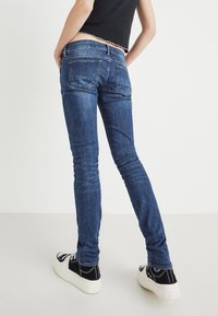 Vaqueros skinny de mezclilla en azul oscuro con un ligero desgaste, diseño de cinco bolsillos y detalles cosidos en la parte trasera y los bolsillos. Combinados con zapatillas negras.
