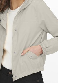 Světle šedá bunda na zip s kapucí a elastickými manžetami. Má dvě boční kapsy a hladký, voděodolný materiál.