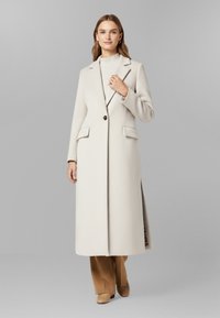 Cappotto lungo di un grigio chiaro, con un design strutturato, rever a punta e un singolo bottone marrone. Tessuto testurizzato e spacchi laterali per facilitare il movimento.