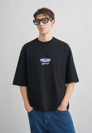 Νέος άνδρας που φοράει μαύρο oversize μπλουζάκι με γραφικό τύπωμα, μπλε τζιν, μαύρα ορθογώνια γυαλιά, στέκεται μπροστά σε απλό φωτεινό φόντο.