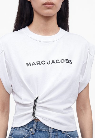Femme portant un T-shirt blanc Marc Jacobs resserré à la taille avec un mousqueton argenté, assorti d’un jean bleu. Partie supérieure du corps montrée.