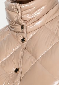 Ljusbeige pufferjacka med en glansig yta, utrustad med hög krage, diamantquiltat mönster och knappar i guldton.