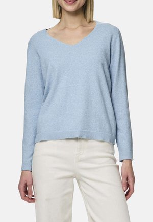 Femme portant un pull à manches longues bleu clair avec un col en V et un pantalon blanc cassé, debout devant un fond uni.