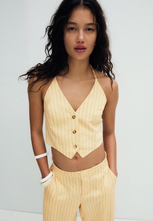 Jonge vrouw met lang, krullend haar, gekleed in een geel gestreept haltertop met knopen en bijpassende broek, handen in de zakken, witte armbanden om de linkerpols.