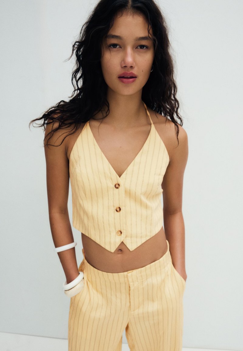 Młoda kobieta o długich kręconych włosach, ubrana w żółty prążkowany top na guziki z wiązaniem na szyi i pasujące spodnie, z rękami w kieszeniach, białe bransoletki na lewym nadgarstku.