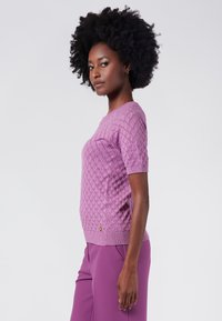 Lyhythihainen, neulottu neulepaita vaaleanliilassa, jossa on teksturoitu timanttikuvio. Ribbaattu helma ja hihansuut, joissa on pieni kultainen logo-aksentti.