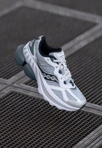 Scarpa da corsa bianca e turchese con tomaia in rete e sintetico, suola spessa e ammortizzata, sistema di allacciatura e dettagli circolari decorativi.