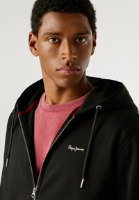 Zwarte zip-up hoodie met trekkoordcapuchon, rode accenten en geborduurd logo. Draagt boven een roze crew neck t-shirt. Gladde stoftextuur.