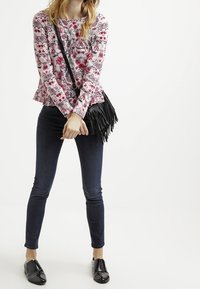 Top de manga larga con estampado floral y puños con volantes, combinado con jeans oscuros de corte ajustado y un bolso de hombro negro con flecos. Zapatos negros tipo slip-on.