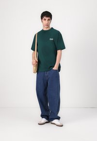 Mørkegrønn bomull t-skjorte med korte ermer, ledsaget av løse blå denimjeans og hvite sneakers, med en enkel logo på brystet.