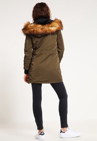 Olivgrön parkasjacka med en plyschfoder huva, dragkedja och tryckknappar, åtdraget midja och sidofickor. Bärs över svarta jeans och sneakers.