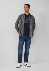 Koszula w kratkę w kolorach granatowym, czerwonym i kremowym na granatowym t-shircie z grafiką, w połączeniu z niebieskimi dżinsami i białymi trampkami z brązowymi akcentami.