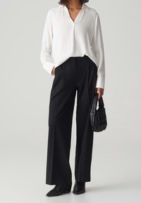 Blouse blanche à manches longues avec un col en V, associée à un pantalon noir taille haute à jambes larges et un petit sac à main noir texturé. Des bottes complètent le look.