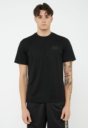 Giovane con capelli mossi che indossa una maglietta nera semplice e pantaloni neri, in piedi su sfondo bianco, che mostra tatuaggi sul braccio sinistro.