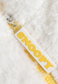 Tela de felpa blanca con un zipper amarillo. El tirador del zipper cuenta con una etiqueta blanca con letras amarillas en relieve que dicen "SNOOPY."