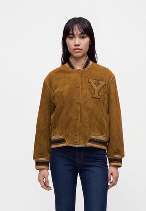 Jeune femme portant une veste varsity en daim marron avec un grand "Y" sur la poitrine et un jean bleu foncé, debout devant un fond uni.
