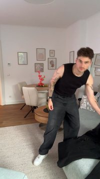 Musta hihaton paita ja löysät mustat farkut valkoisilla tennareilla. Vaaleanharmaa sohva ja kuvioidut tyynyt modernissa, hyvin valaistussa huoneessa.