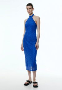 Blaue Spitzen-Halskappendresse mit passender Silhouette, knielang und mit komplexer Spitzenstruktur. Kombiniert mit schwarzen Plateausandalen.