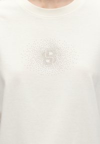 T-shirt blanc avec un motif circulaire de petits clous formant le chiffre 5 sur la poitrine.