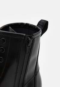 Close-up af sort læderstøvle med sidesyning, lynlås, snørebånd, syninger og stofstrop øverst.