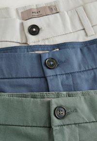 Pile de trois pantalons pliés en beige, bleu et vert, chacun avec une fermeture à bouton noir à la taille.