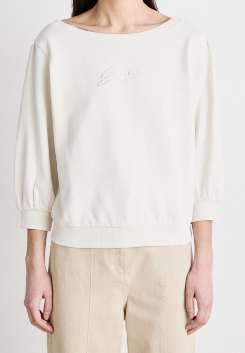 Femme portant une chemise blanche à manches longues avec un large décolleté et un pantalon beige, debout devant un fond uni.