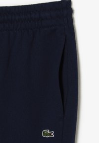 Marineblå joggebukser laget av mykt stoff, med elastisk linning og sidelommer. Inkluderer en grønn og hvit brodert logo.