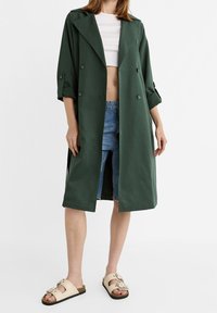 Długi zielony płaszcz w lekkim materiale, podwójnie zapinany z dużymi guzikami, z podwiniętymi rękawami, w zestawieniu z dżinsowymi szortami i sandałami.