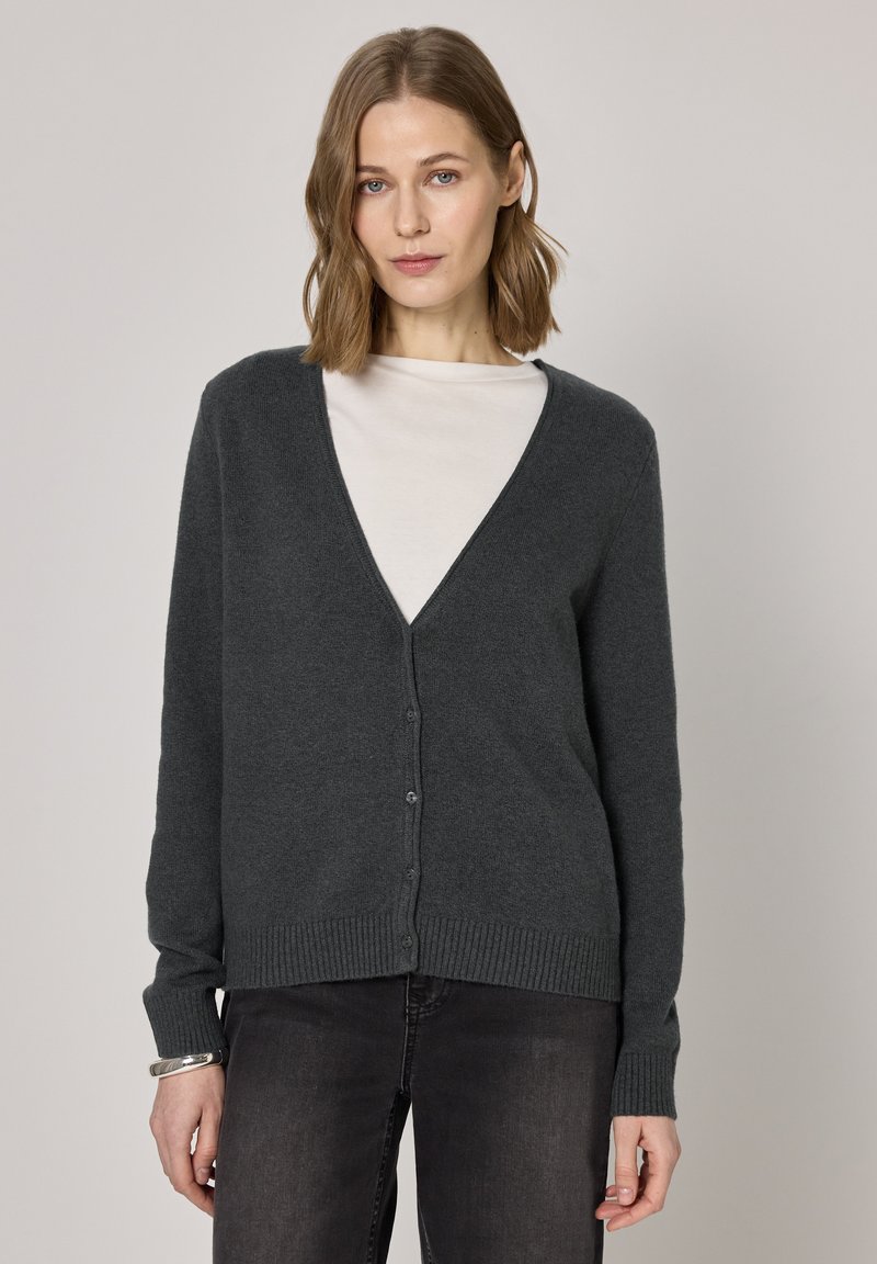 Cardigan gris en maille avec un col en V, fermeture à boutons, poignets et ourlet côtelés. Porté sur un haut blanc, il présente une texture douce et une coupe décontractée.