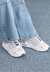 Baskets athlétiques blanches et beiges avec des accents en mesh et en daim, présentant une semelle épaisse, une tige texturée et un design à lacets.