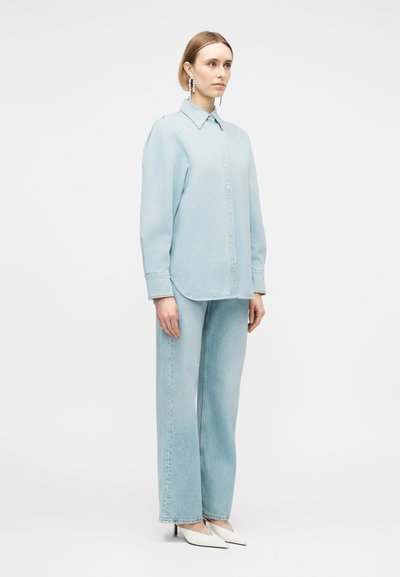 Γυναίκα που στέκεται φορώντας ανοιχτό γαλάζιο πουκάμισο τζιν και τζιν παντελόνι με φαρδιά μπατζάκια, λευκές μυτερές γόβες και μεγάλα κρεμαστά σκουλαρίκια, ουδέτερο φόντο.