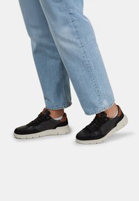 Mustad tossud, millel on sile nahk ja sametine viimistlus, valge kummist põhi ja pruuni nahast detailid kandade juures, koos helesiniste teksadega.