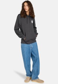 Sudadera gris con capucha, bolsillo frontal y cordones. Combinada con jeans de mezclilla ligeros y holgados de color azul claro y calzado beige.