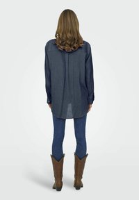 Camicia blu scuro a maniche lunghe con orlo arrotondato e texture sottile, abbinata a jeans aderenti e stivali da cowboy marroni.