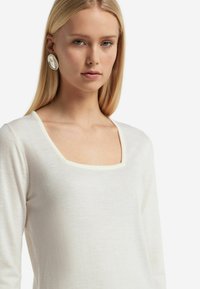 Vit långärmad topp med fyrkantig halsringning och ljus kontrasterande kant, tillverkad av mjukt tyg. Modellen bär stora runda örhängen.