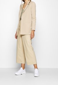 Beige maßgeschneiderter Blazer mit einem Knopf, kombiniert mit weiten beigen Hosen und weißen sportlichen Sneakern. Glattes Material, minimalistisches Design.