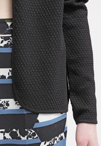 Svart texturerad jacka med en åtsittande design, med rundade kanter och en kontrasterande mönstrad kjol under, inklusive blommiga och randiga detaljer.