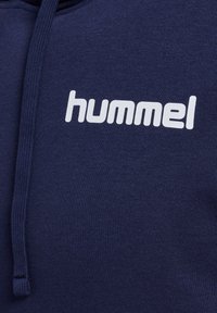 Mörkblå hoodie med en texturerad tygkvalitet. Har en vit "hummel"-logotyp på bröstområdet och en huva med dragsko.