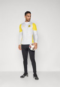 Světle šedé sportovní tričko s dlouhým rukávem a žlutými detaily, s černým logem. Černé slim-fit kalhoty a černé fotbalové boty. Drží fotbal.