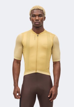 Atletisk mand iført gul tekstureret cykeltrøje med korte ærmer og tætsiddende brune cykelshorts, stående med hænder afslappet ned langs siderne.