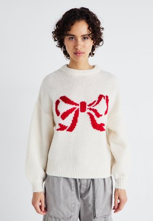 Pull en tricot blanc avec un grand motif de nœud rouge sur le devant. Encolure ronde et poignets côtelés ajoutent de la texture.