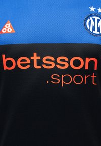 Η ποδοσφαιρική φανέλα της Ίντερ Μιλάνο με μπλε και μαύρες περιοχές, λογότυπο χορηγού Betsson.sport σε πορτοκαλί και σήματα ACG και συλλόγου κοντά στον γιακά.