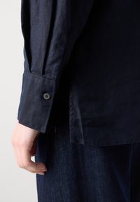 Chemise à manches longues bleu marine avec un poignet à bouton, texture douce et fente sur le côté, associée à un pantalon en denim foncé.