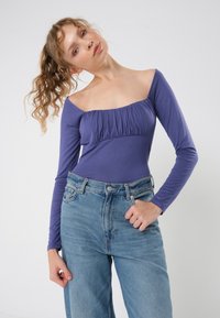 Lilla langermet topp med samlet bust, kombinert med høyt liv blå jeans. Tøyet er glatt, og designet har en av-skulderen utringning.