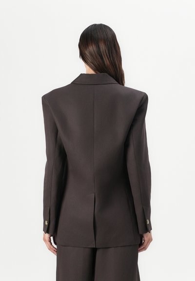 Blazer marrone su misura con silhouette strutturata, spalle accentuate e uno spacco sul retro. Include polsini con bottoni e tessuto morbido.