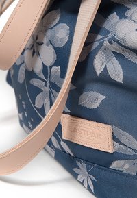 Blue fabric bag with white floral pattern, featuring a smooth pink leather handle and a small embossed label with the brand name.