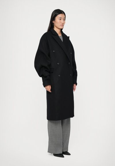 Femeie în picioare, purtând o haină neagră oversized cu două rânduri de nasturi, pantaloni gri largi și pantofi negri cu vârf ascuțit, pe un fundal simplu.
