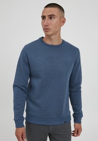 Puha szövetből készült kék pulóver, bordázott kerek nyakkal és mandzsettákkal, a mellkasán egy diszkrét logó részlettel.