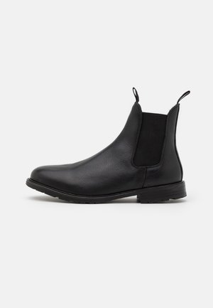 Stivale Chelsea in pelle nera con pannelli laterali elastici, linguette anteriori e posteriori e tacco basso a blocco su sfondo bianco.