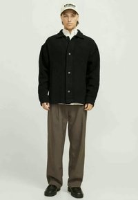 Manteau noir à boutons, texture côtelée, coupe surdimensionnée, porté sur une chemise blanche. Un pantalon marron et des chaussures noires complètent la tenue, avec une casquette.